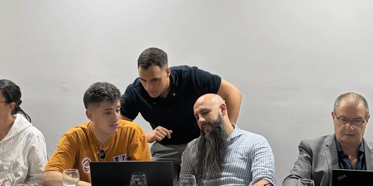 Un integrante del equipo, parado detrás de la mesa e inclinado sobre una notebook, señalando algo en la pantalla mientras los demás miran.